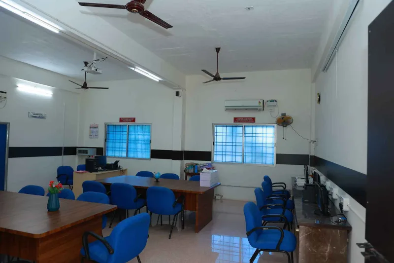 A seminar room set out with blue chairs around tables, a ceiling projector, and computers along the side wall.